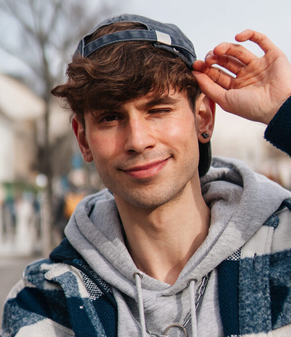 Kostas Kind, Content Creator und Autor, steht draußen in lässiger Kleidung und zwinkert der Kamera zu. Er trägt eine blau-graue Karojacke über einem grauen Hoodie und eine umgedrehte Basecap, während er mit einer Hand seinen Cap-Schirm festhält. Im Hintergrund ist eine unscharfe Stadtumgebung zu erkennen.
