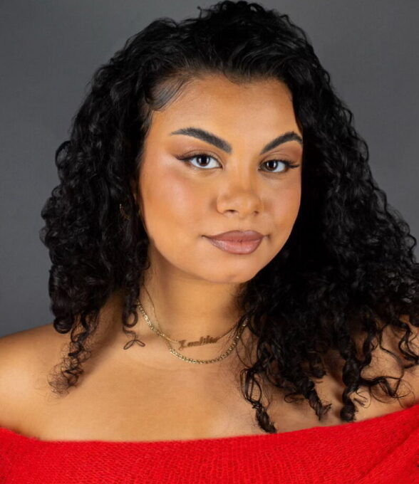 Laetitia Colacicco, Content Creatorin und Moderatorin, blickt mit ruhigem, selbstbewusstem Ausdruck in die Kamera. Sie trägt ein leuchtend rotes, schulterfreies Oberteil und auffälliges Make-up; ihre dunklen, lockigen Haare fallen ihr über die Schultern. Der Hintergrund ist neutral grau gehalten.