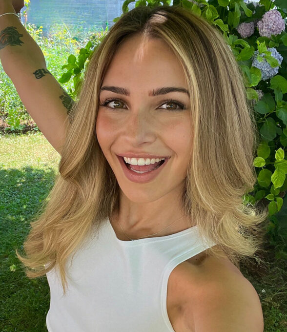 Lenaslogik, eine Content Creator und ehemalige Polizistin, trägt ein weißes Oberteil und hat lange, wellige Haare. Sie hebt einen Arm in die Luft und steht vor einer grünen Pflanzenkulisse.