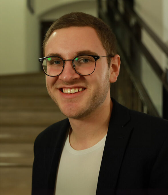 Maik Jahn trägt ein schwarzes Sakko und ein helles T-Shirt. Er steht auf einer Treppe in einem Innenraum.