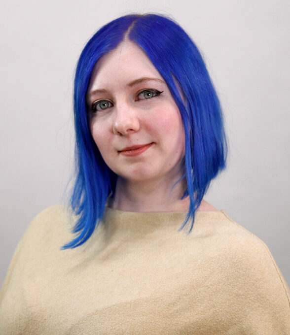 Marylin Marx mit blauen Haaren, trägt ein beiges Oberteil. Der Blick ist neutral, der Kopf leicht zur Seite geneigt.