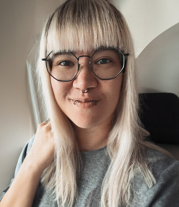 Mira Minks mit langen, blonden Haaren trägt ein graues Sweatshirt und sitzt in einem Flugzeug. Sie hat eine Hand an ihrem Hals.