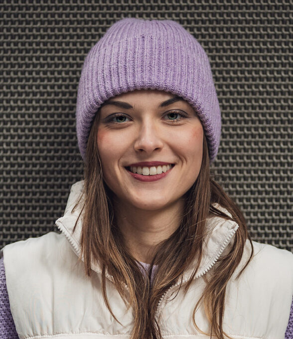 Nerdzika trägt eine lila Mütze und eine weiße Weste. Die Person hat lange, braune Haare und steht vor einem strukturierten Hintergrund.