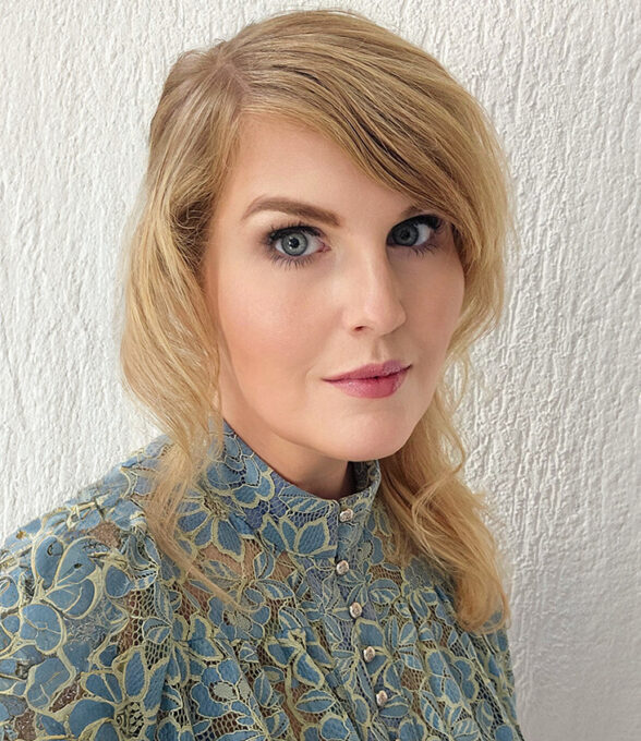 Nina Schild blickt mit neutralem, leicht ernstem Ausdruck in die Kamera. Sie hat lange, blonde Haare und trägt ein blau-grünes, gemustertes Oberteil. Der Hintergrund ist eine weiße, strukturierte Wand.