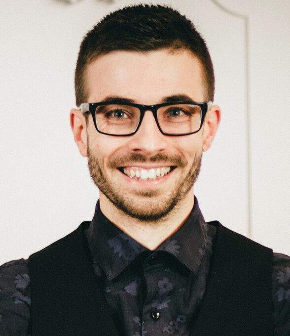 Pascal Simon lächelt freundlich in die Kamera. Er trägt eine dunkle Brille, ein schwarzes Hemd mit dezentem Muster und eine schwarze Weste. Der Hintergrund ist hell und schlicht, was den Fokus ganz auf sein Gesicht lenkt.