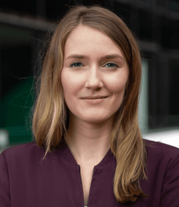 Vanessa Bitter, Chief Operating Officer, mit langen, blonden Haaren, trägt ein lila Oberteil. Der Hintergrund zeigt unscharfe architektonische Elemente.