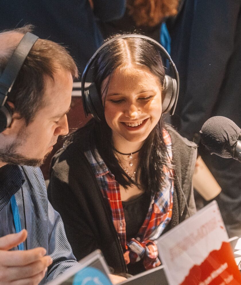 Zwei Personen mit Kopfhörern sitzen zusammen vor Mikrofonen und lachen.