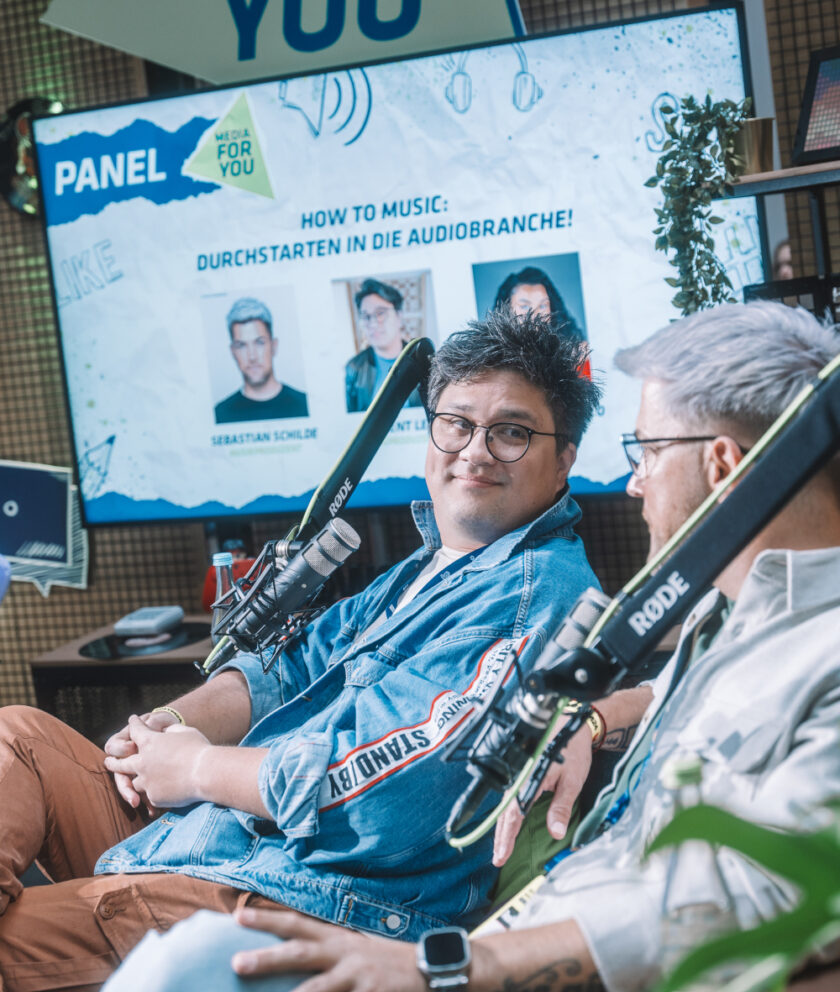 Zwei Männer sitzen mit Mikrofonen in einer Panel-Diskussion, hinter ihnen ein Bildschirm mit dem Thema „How to Music: Durchstarten in die Audiobranche".