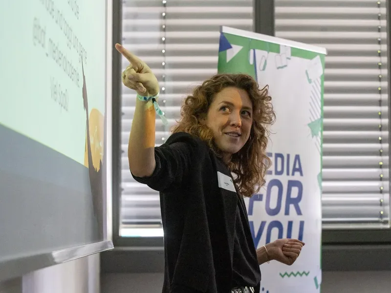 Frau mit lockigem Haar zeigt auf eine Wand oder Leinwand und lächelt, vor einem Banner mit der Aufschrift "MEDIA FOR YOU".