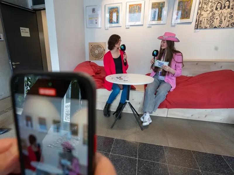 Gerlinde trägt einen pinken Hut und sitzt an einem Tisch mit einer anderen Person. Beide halten Mikrofone. Im Hintergrund sind Bilder an der Wand und eine rote Couch.