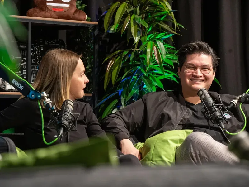 Das Bild zeigt zwei Personen, die auf einem Sofa sitzen und sich unterhalten. Beide haben ein Mikrofon vor sich, es sieht nach einer Gesprächssituation oder einem Interview aus. Im Hintergrund sind Pflanzen zu sehen.