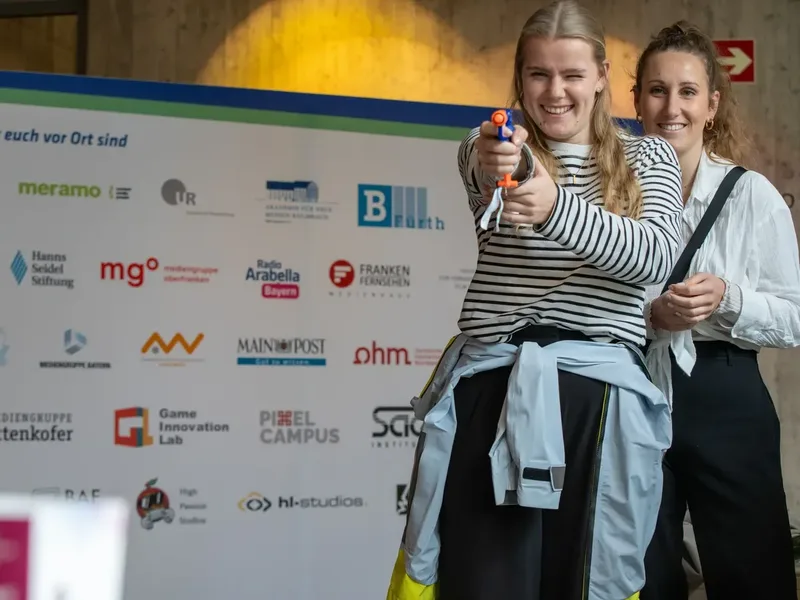 Zwei junge Frauen stehen lachend vor einer Sponsorenwand, während eine von ihnen mit einer Spielzeugpistole zielt.