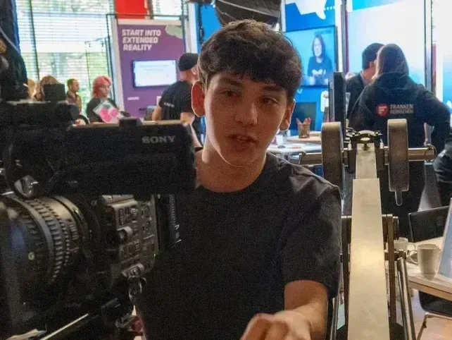 Ein junger Mann sitzt hinter einer großen Kamera auf einem Stativ und blickt konzentriert nach vorne. Im Hintergrund sind weitere Personen und Ausrüstung zu sehen.