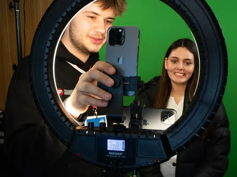 Zwei Personen stehen vor einem grünen Hintergrund. Ein junger Mann hält ein Smartphone vor eine Ringleuchte, und eine junge Frau lächelt in die Kamera.