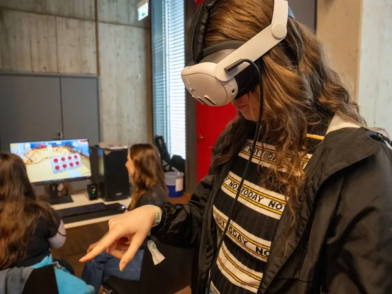 Person mit Virtual-Reality-Brille zeigt mit dem Finger nach unten, im Hintergrund zwei Personen vor einem Bildschirm.