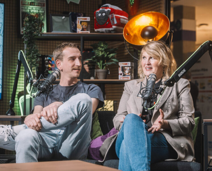 Zwei Personen sitzen auf einer Couch vor Mikrofonen und sprechen angeregt in einer Talkrunde, während sie aufgenommen werden.