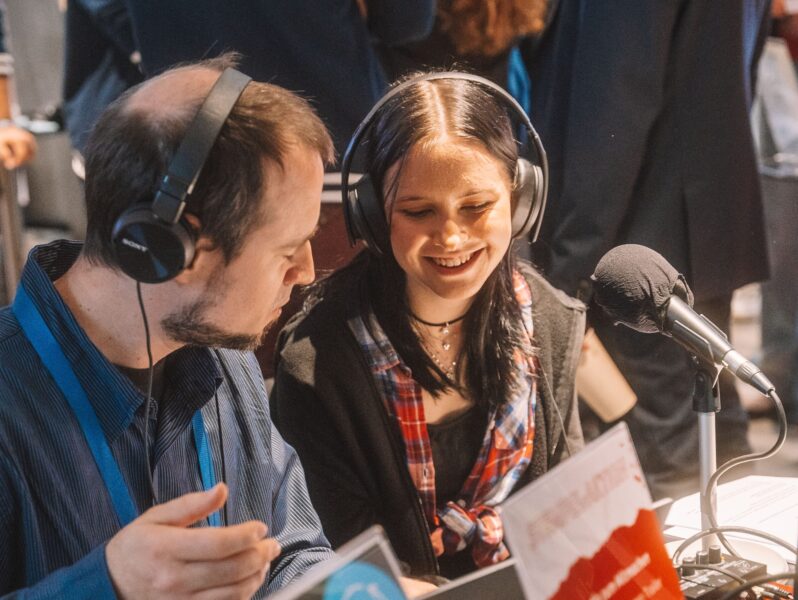 Zwei Personen mit Kopfhörern sitzen zusammen vor Mikrofonen und lachen.