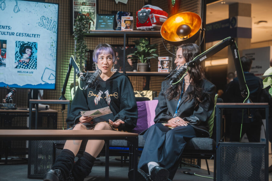 Zwei Frauen sitzen auf einer Bühne mit Mikrofonen vor sich und diskutieren, im Hintergrund ein Bildschirm mit dem Namen „Melissa Lee“.