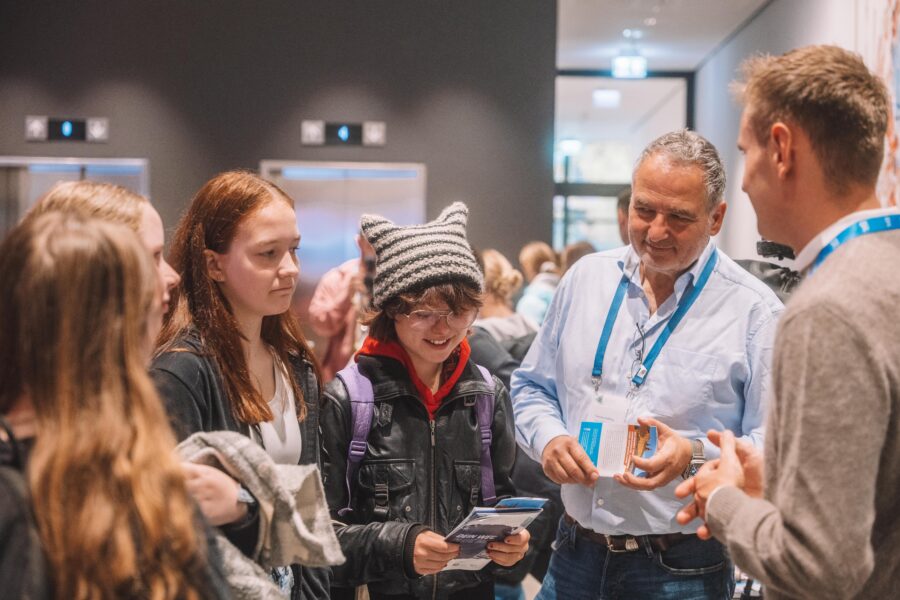 Eine Gruppe von Menschen steht zusammen, darunter zwei junge Frauen, ein Kind mit Mütze und zwei erwachsene Männer. Sie unterhalten sich und schauen auf ein Buch oder eine Broschüre.