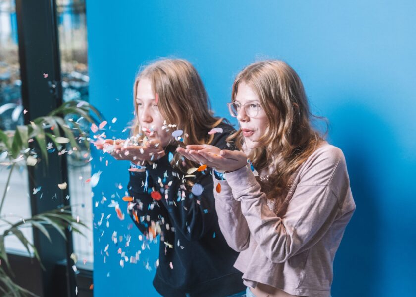 Zwei Mädchen stehen vor einer blauen Wand und blasen buntes Konfetti in die Luft.