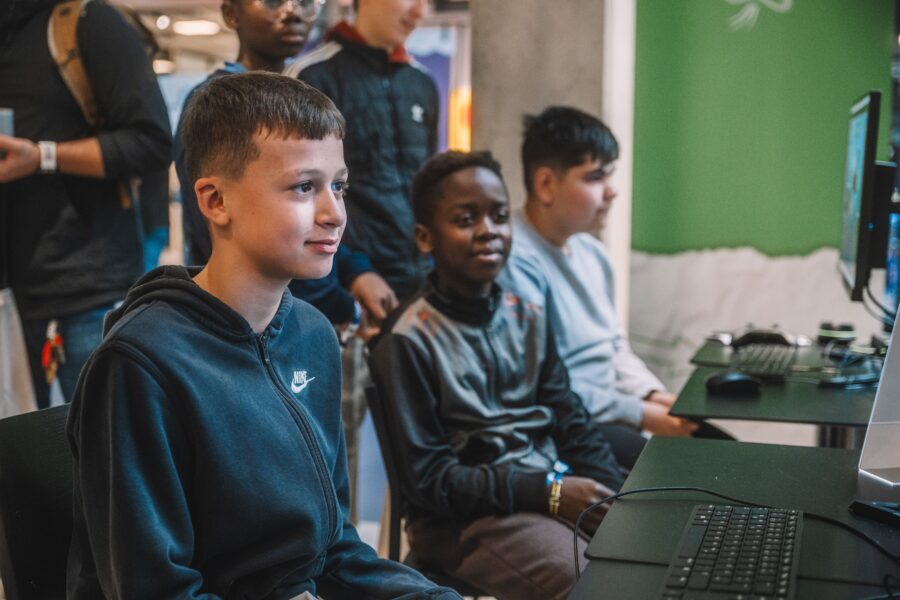 Drei Jungen sitzen nebeneinander an Computern und schauen konzentriert auf die Bildschirme. Im Hintergrund sind weitere Personen zu sehen.