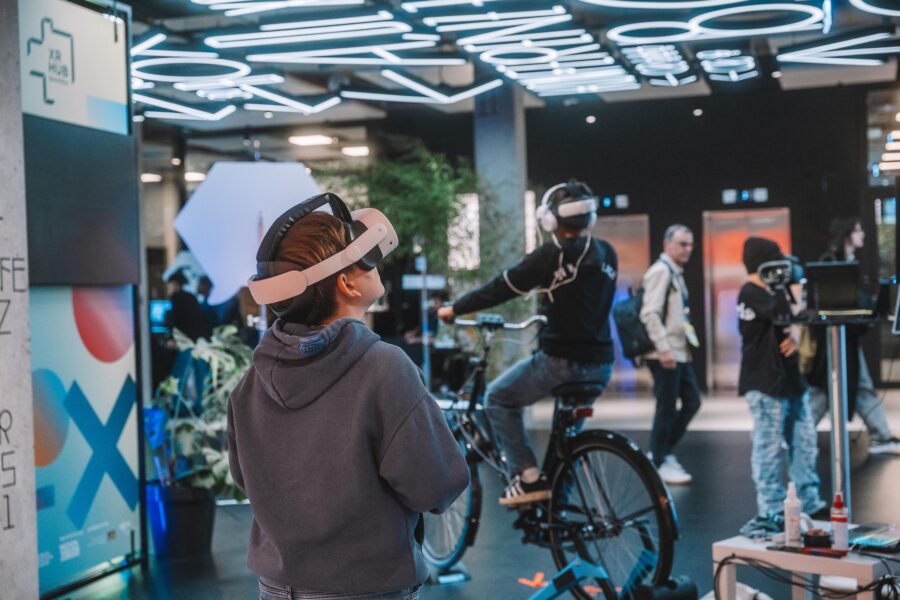 Mehrere Personen tragen VR-Brillen. Eine Person sitzt auf einem Fahrrad und nutzt die VR, während andere stehen und zuschauen. Die Szene spielt in einem modernen, gut beleuchteten Raum.