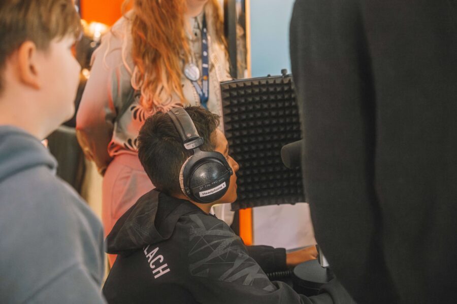 Mehrere Personen sind in einem Raum zu sehen, einer sitzt vor einem Mikrofon mit Kopfhörern und einem Schallschutzschild, andere stehen und beobachten.