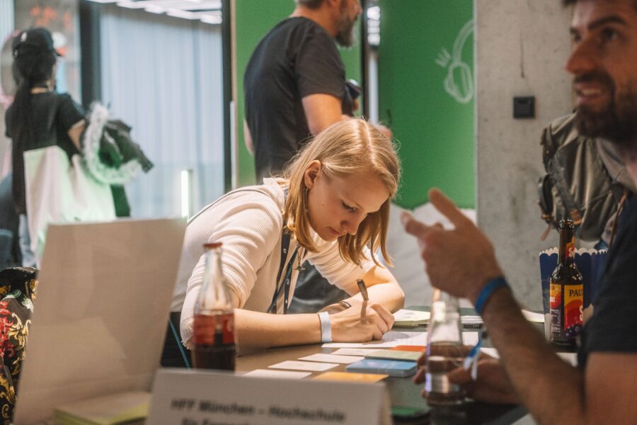 Eine junge Frau sitzt an einem Tisch und schreibt konzentriert auf kleine Zettel. Vor ihr steht eine Cola-Flasche, im Vordergrund ist ein Mann mit Bart und Getränk unscharf zu sehen. Im Hintergrund sind weitere Personen und ein grüner Raum zu erkennen.