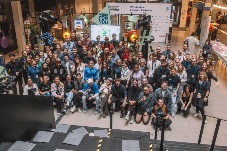 Gruppenfoto vieler Mitarbeitender von „Media for You“ in einer Halle, alle blicken in die Kamera und lächeln.