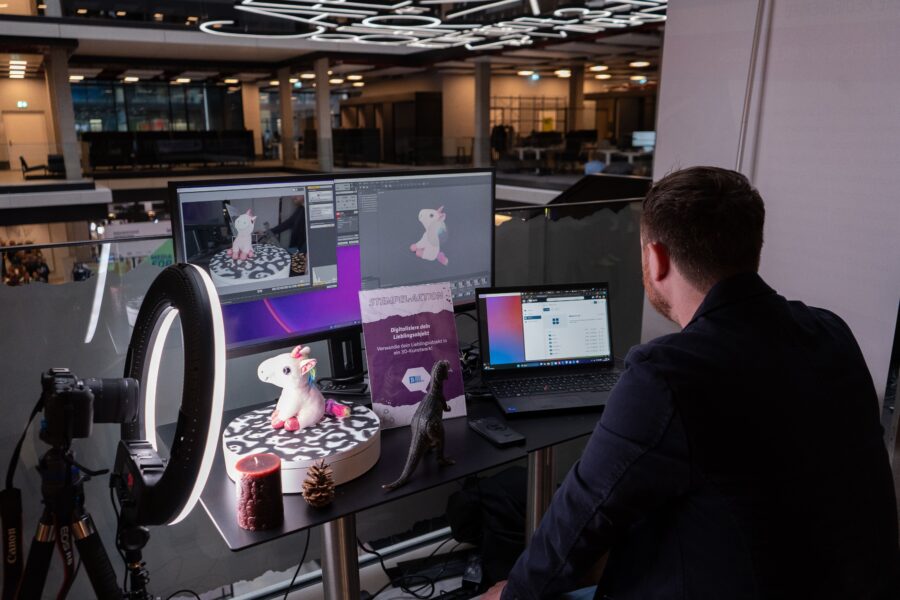 Mann arbeitet an einem Stehtisch mit Laptop und zwei Monitoren, auf denen eine 3D-Einhorn-Figur zu sehen ist. Daneben stehen eine Ringlichtlampe, Kamera und kleine Dekorationen. Im Hintergrund ein modernes Büro.