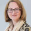 Anette Kolb mit schulterlangen, blonden Haaren, trägt einen hellen Schal und eine Bluse mit floralem Muster. Sie schaut leicht zur Seite.