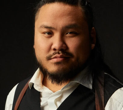 Das Portrait eines asiatisch gelesenen Mannes mit Bart und zu einem Zopf gebundenen Haaren. Er trägt ein weißes Hemd mit braunen Hosenträgern und eine schwarze Weste.