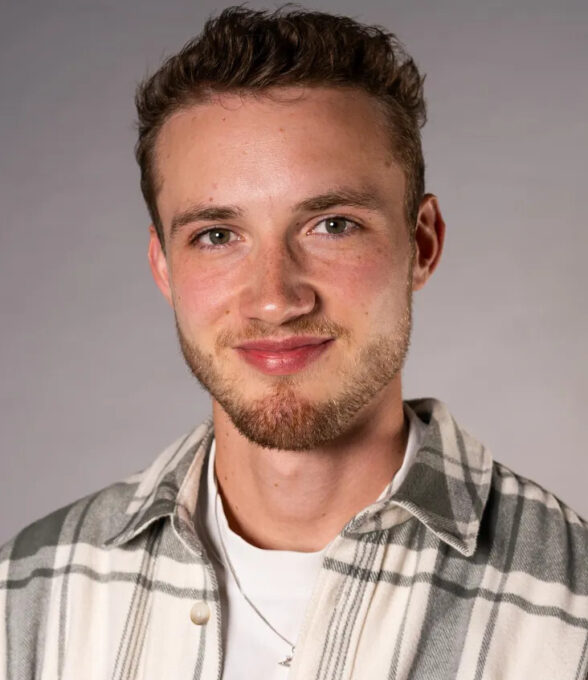 Portrait eines jungen Mannen mit braunem Bart und kurzen Haaren, der ein weißes Shirt und darüber ein Karohemd trägt.