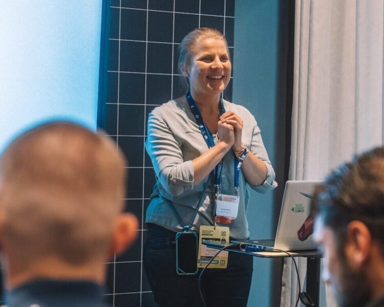 Eine Frau hält einen Vortrag vor einer kleinen Gruppe. Sie steht lächelnd vor einem Laptop und einer Präsentationsfläche, trägt ein Namensschild und spricht zu den Teilnehmenden, die im Vordergrund sitzen.