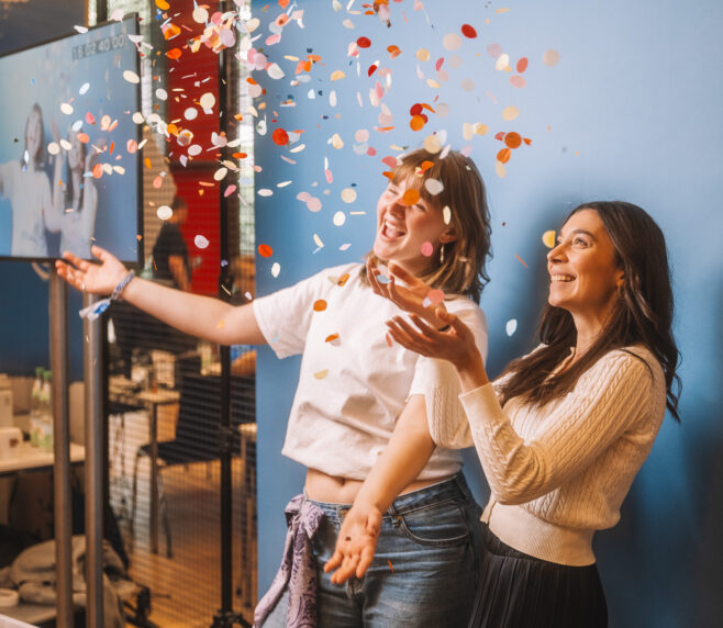 Zwei junge Frauen sind vor einem blauen Hintergrund zu sehen. Sie lachen, während sie Confetti für eine slow motion Aufnahme in die Luft werfen.