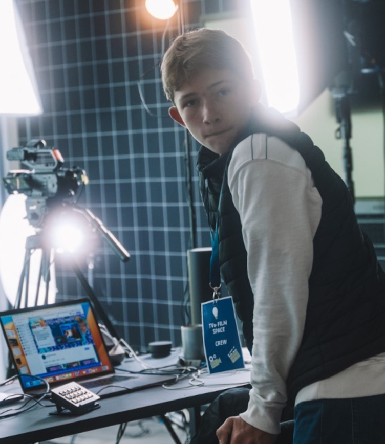 Eine Person steht an einem Tisch mit einem Laptop und mehreren Kameras. Helle Scheinwerfer beleuchten die Szene. Die Person trägt eine dunkle Weste und hat einen Ausweis um den Hals.