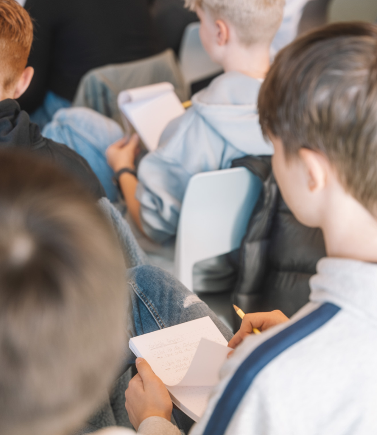 Gruppe von Jugendlichen, die in einem Workshop sitzen. Einige halten Notizblöcke und Stifte. Die Kleidung ist leger, mit verschiedenen Farben und Stilen.