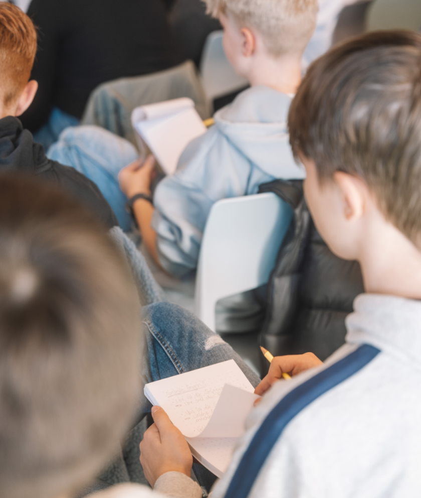 Gruppe von Jugendlichen, die in einem Workshop sitzen. Einige halten Notizblöcke und Stifte. Die Kleidung ist leger, mit verschiedenen Farben und Stilen.