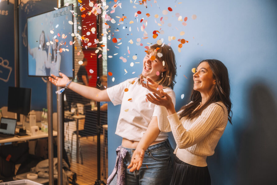 Zwei junge Frauen sind vor einem blauen Hintergrund zu sehen. Sie lachen, während sie Confetti für eine slow motion Aufnahme in die Luft werfen.