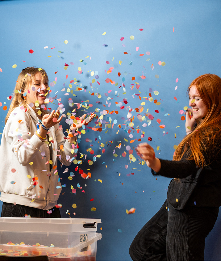 Zwei junge Frauen lachen und werfen buntes Konfetti in die Luft vor einer blauen Wand.