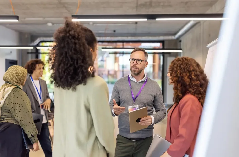 Eine Gruppe von fünf Personen unterhält sich bei einer Networking-Veranstaltung in einem modernen Bürogebäude, ein Mann mit Tablet spricht mit zwei Frauen im Vordergrund.