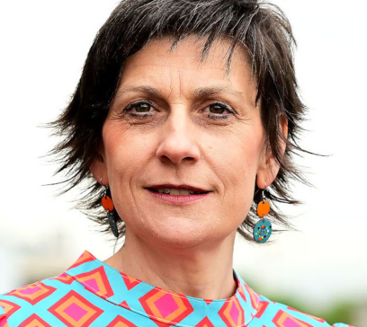 Porträt einer Frau mit kurzem dunklem Haar, die bunte Ohrringe trägt. Sie hat eine bunte Bluse mit geometrischem Muster an und blickt direkt in die Kamera. Der Hintergrund ist verschwommen und hell.