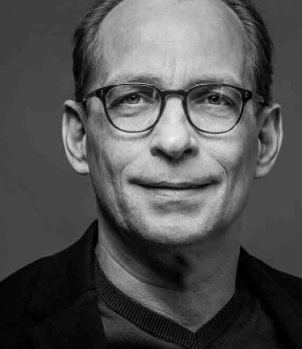 Schwarzweiß-Porträt eines älteren Mannes mit kurzen Haaren und Brille, der in die Kamera lächelt. Er trägt ein dunkles Oberteil und einen Blazer.