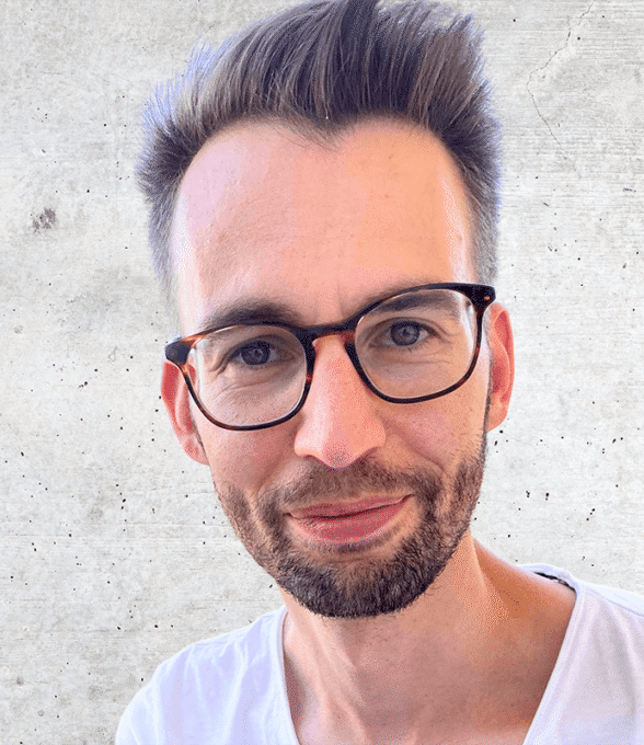 Das ist ein Porträtfoto eines jungen Mannes mit Brille, kurzen, nach oben gestylten Haaren und einem kurzen Bart. Er trägt ein weißes T-Shirt und lächelt leicht. Der Hintergrund ist eine graue Wand.