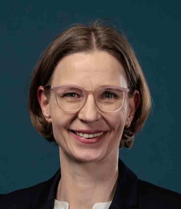 Porträtfoto einer lächelnden Frau mit kurzen braunen Haaren und durchsichtiger Brille. Sie trägt ein schwarzes Sakko und ein weißes Oberteil. Der Hintergrund ist dunkelblau.