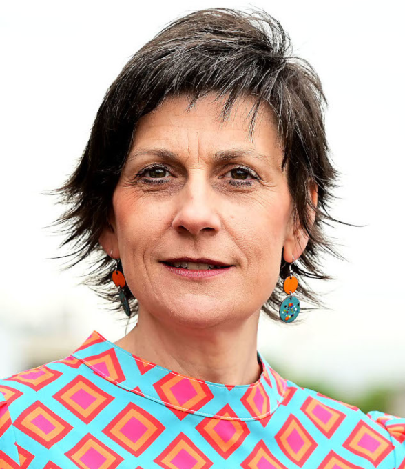 Porträt einer Frau mit kurzem dunklem Haar, die bunte Ohrringe trägt. Sie hat eine bunte Bluse mit geometrischem Muster an und blickt direkt in die Kamera. Der Hintergrund ist verschwommen und hell.